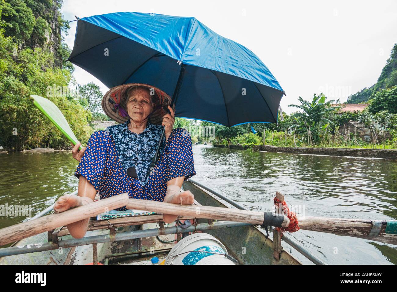 An elderly woman uses her feet to push the oars of a rowboat, while holding a fan and umbrella, taking tourists down the Ngo Dong River, Vietnam Stock Photo