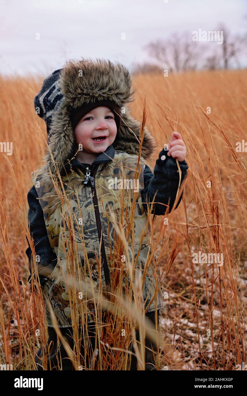 little boy wearing camoflauge vest and  fur-lined trapper hat in a country field Stock Photo