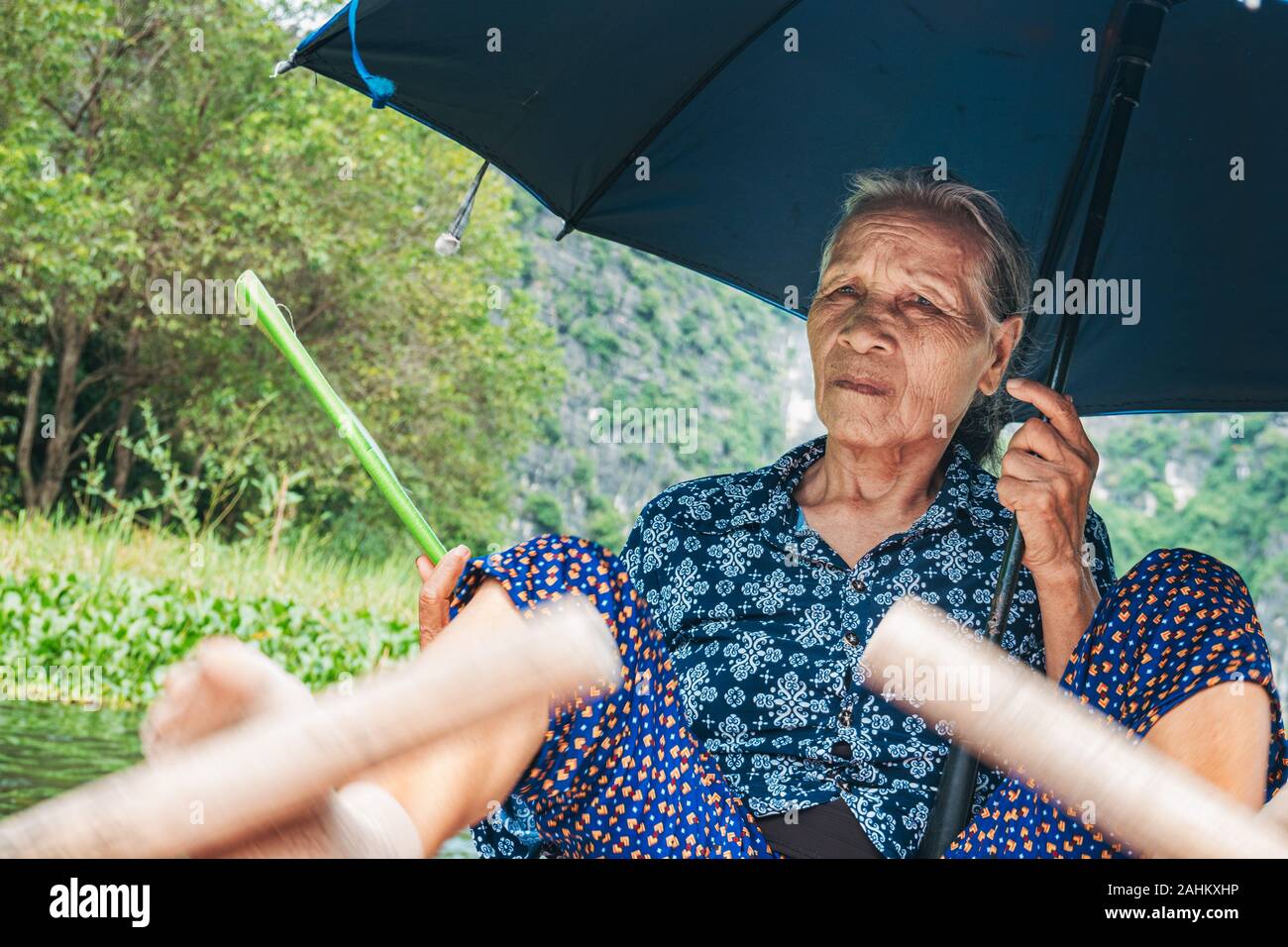 An elderly woman uses her feet to push the oars of a rowboat, while holding a fan and umbrella, taking tourists down the Ngo Dong River, Vietnam Stock Photo