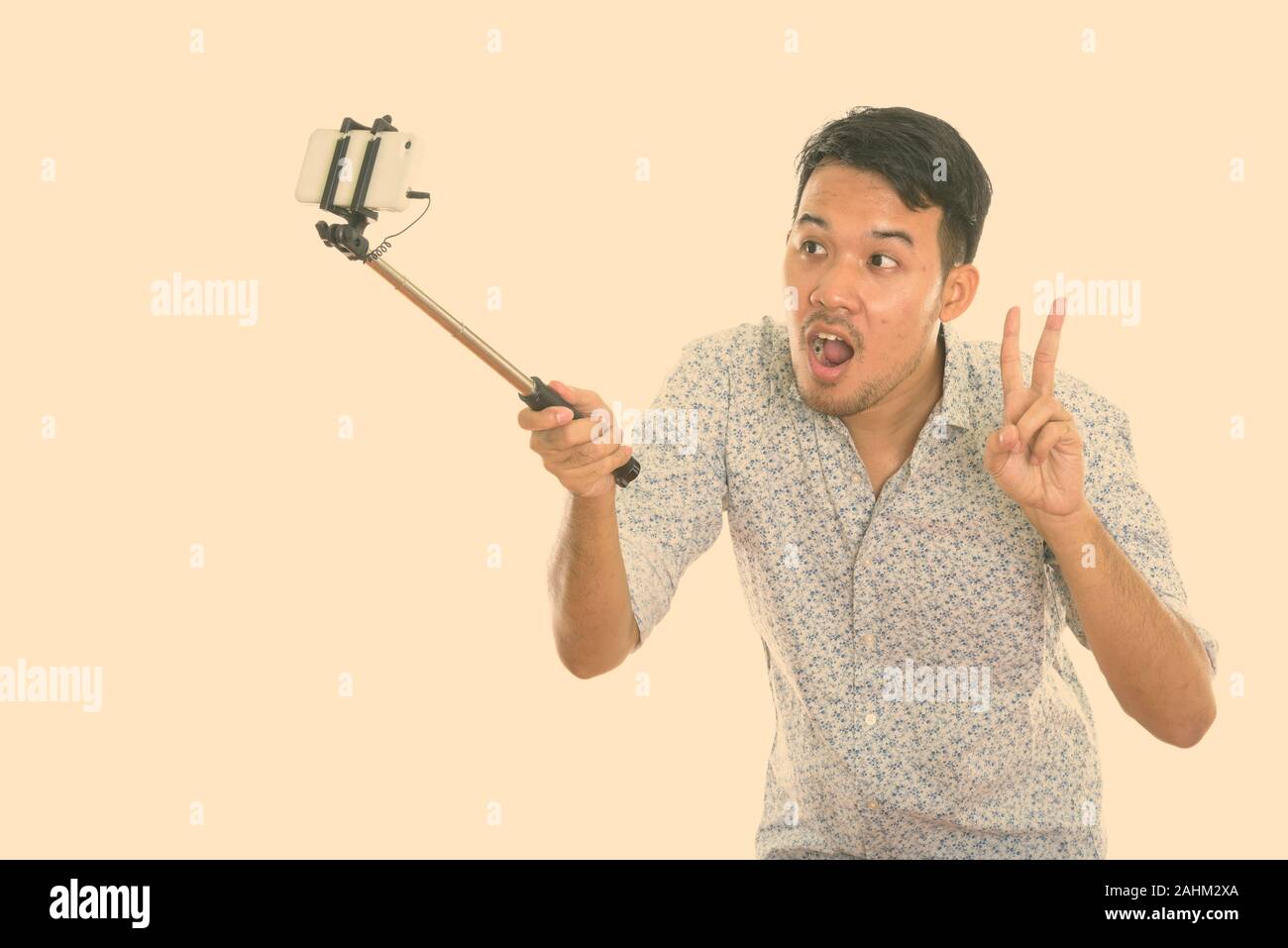 Young Asian man giving peace sign while taking selfie picture with mobile  phone and holding selfie stick Stock Photo - Alamy