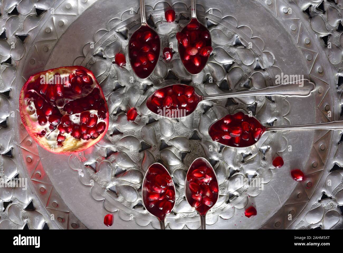 An old ornate plate made of tin with silver spoons holding pomegranate seeds along with half a pomegranate Stock Photo