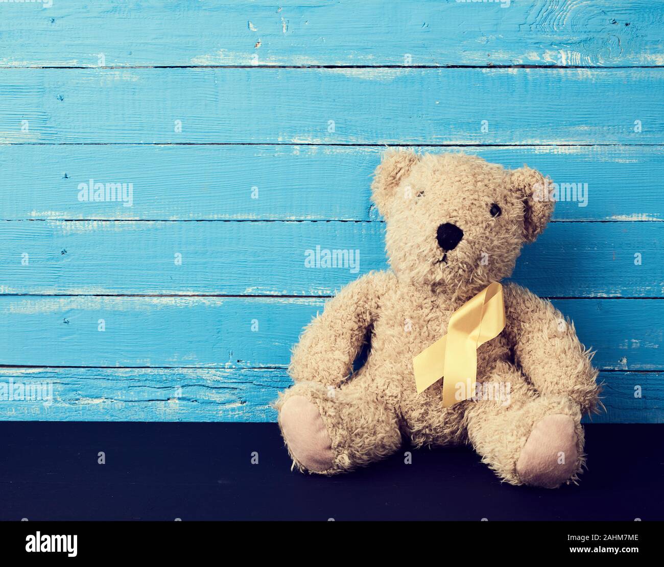 brown teddy bear sits and a yellow silk ribbon on a blue wooden background, concept of the fight against childhood cancer. problem of suicides and the Stock Photo