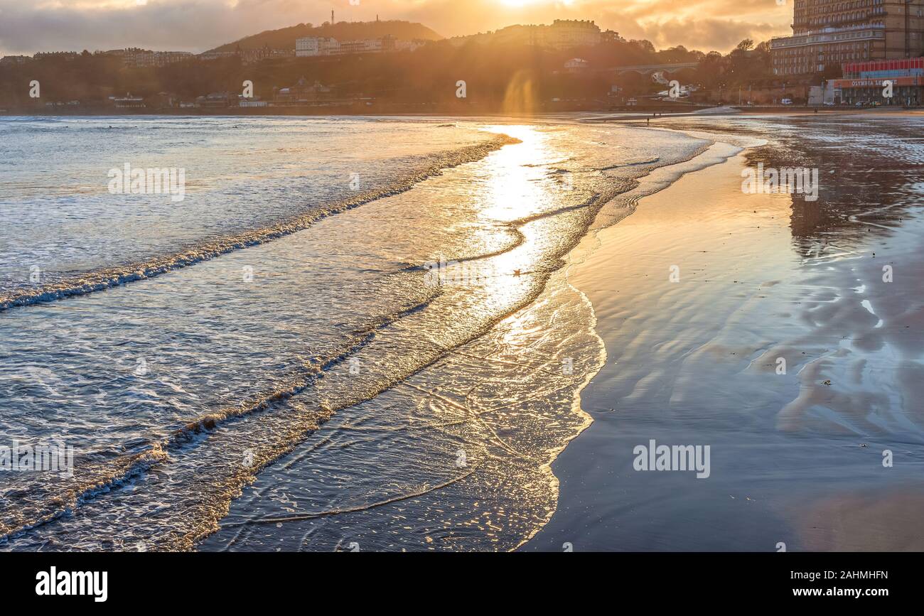 South Beach at Scarborough looking towards the sun.  Waves lap onto the shore with buildings reflected in the wet sand. Hills are in the distance. Stock Photo