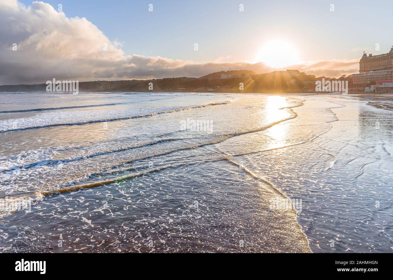 South Beach at Scarborough looking towards the sun.  Waves lap onto the shore with buildings reflected in the wet sand. Hills are in the distance. Stock Photo