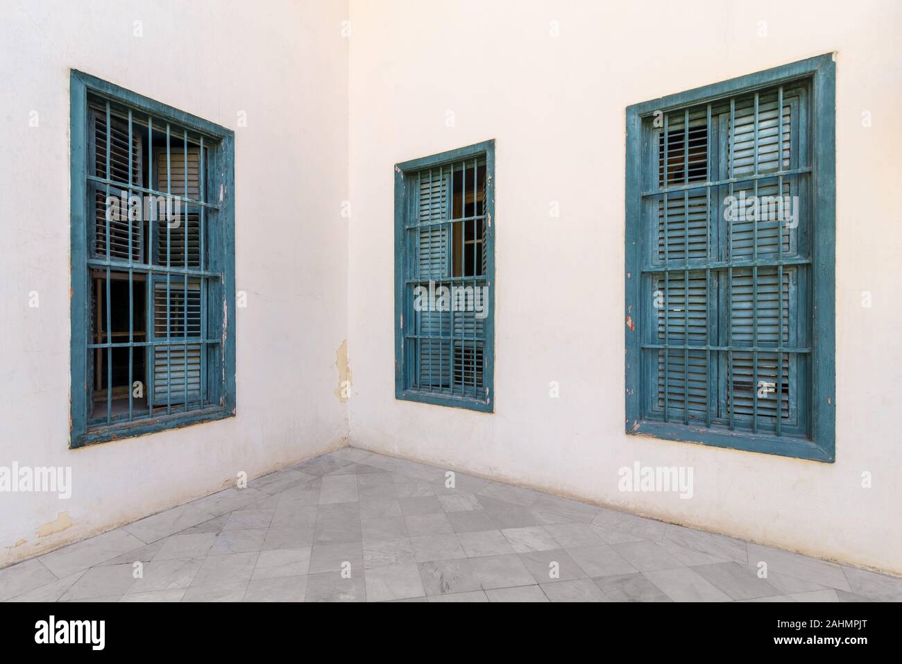 Corner white wall with three grunge windows with wooden green shutters and wrought iron bars and tiled white marble floor Stock Photo