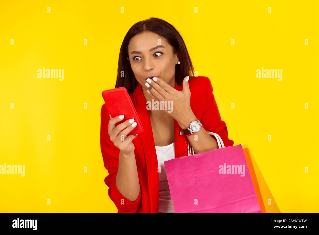 Young beautiful woman holding cellphone in hand examining it with surprised face, big eyes covering mouth with hand isolated on yellow backdrop. Great Stock Photo