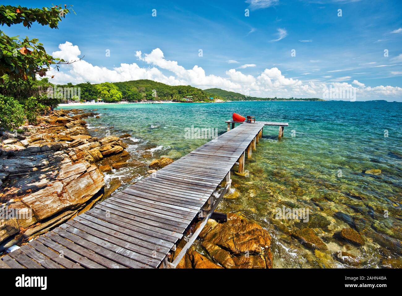 Wooden jetty in tropical beach, textured stones of the coastline is at foreground, and eastern coastline of Ko Samet island is at background, Thailand Stock Photo