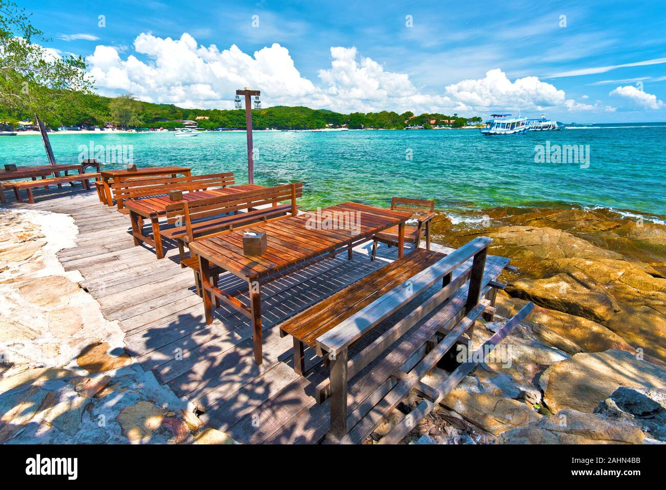 Beach restaurant wooden tables in the border of sea, textured stones of the coastline is at foreground, and eastern coastline of Ko Samet island is at Stock Photo