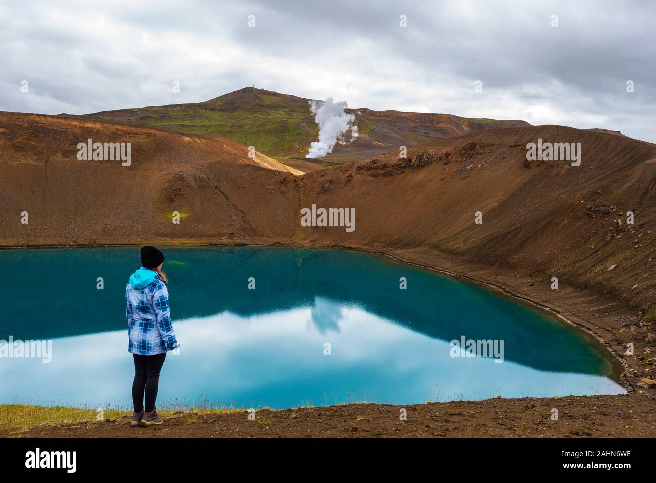 Girl teenager is staying in the border of Krafla volcanic crater and Viti lake. Vapor of geothermal power plant unit is at background, Nordurland eyst Stock Photo
