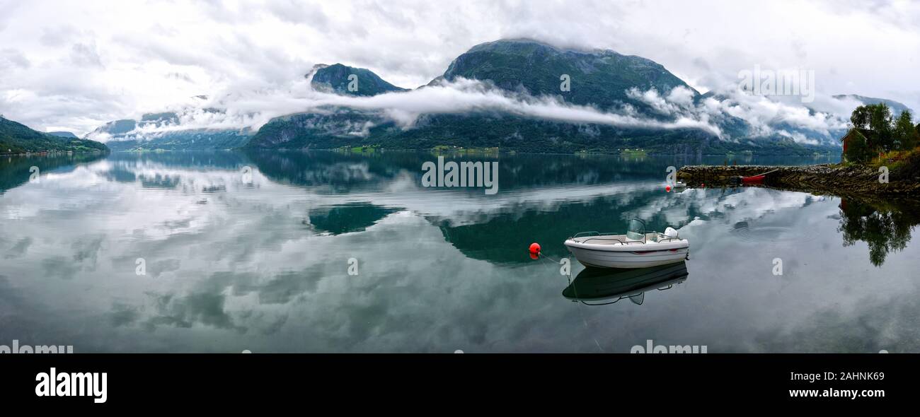 Panoramic view of Lustrafjorden covered with mist, mountains and foggy pattern is reflecting in the water, white boat and  lonely hut in the border of Stock Photo