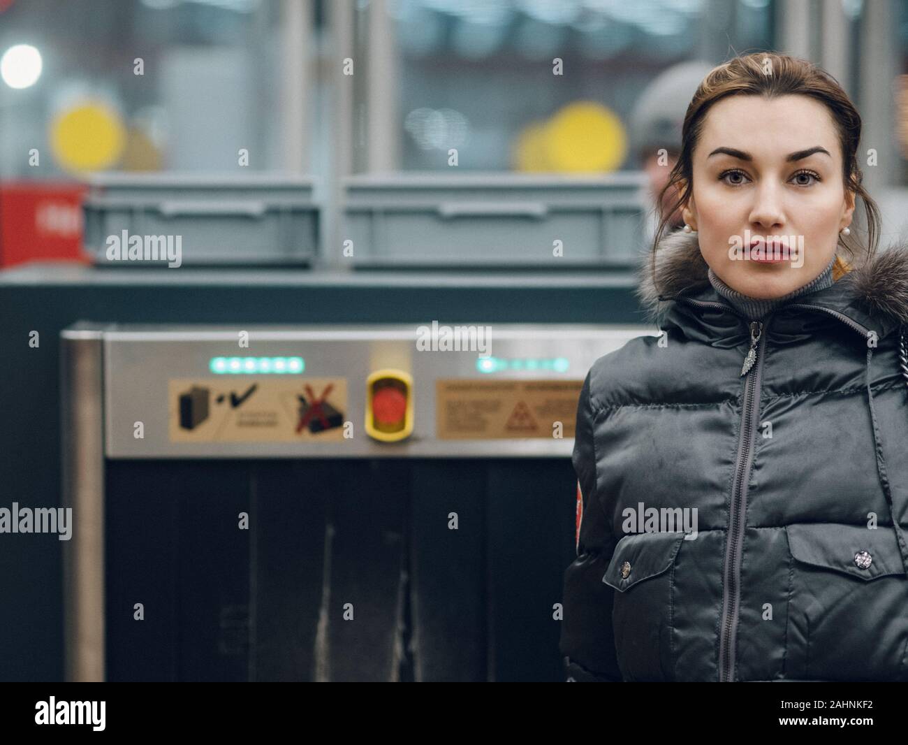 A young woman is looking at the camera. A brunette in demi-season clothes is standing near the security counter in the subway. In the background is a Stock Photo