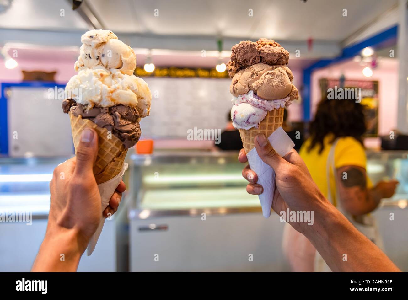 A close up and first person perspective of a man holding two waffles cone with scoops of ice cream at a shop, with blurry menu in the background Stock Photo