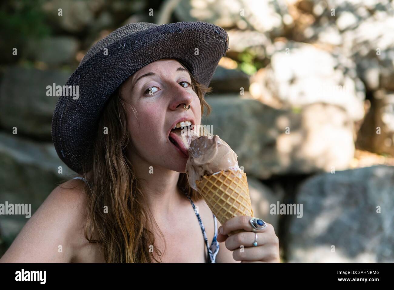 A close up and front portrait of a caucasian with pierced nose licking an ice cream cone. sitting in the shade near some blurry rocks Stock Photo