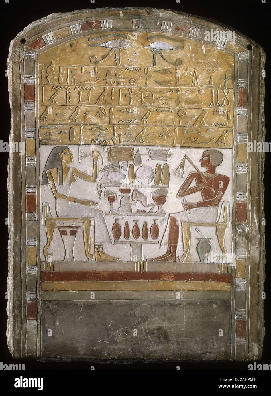 Ancient Egyptian. Stela of Amenemhat and Yatu. 1870 BC–1770 BC. Egypt. Limestone and pigment This stela, or decorated stone monument, depicts a man named Amenemhat seated across from his mother Yatu. A table covered in offerings including bread, meat, and drinks separates them. Amenemhat is portrayed holding a flywhisk, a sign of authority. A blue stone jar designed to hold kohl (eye paint) with an applicator sticking out of its top is shown under his chair. An ointment container is depicted beneath the chair of Yatu, who holds a lotus blossom to her nose. In ancient Egypt, the lotus was a sym Stock Photo