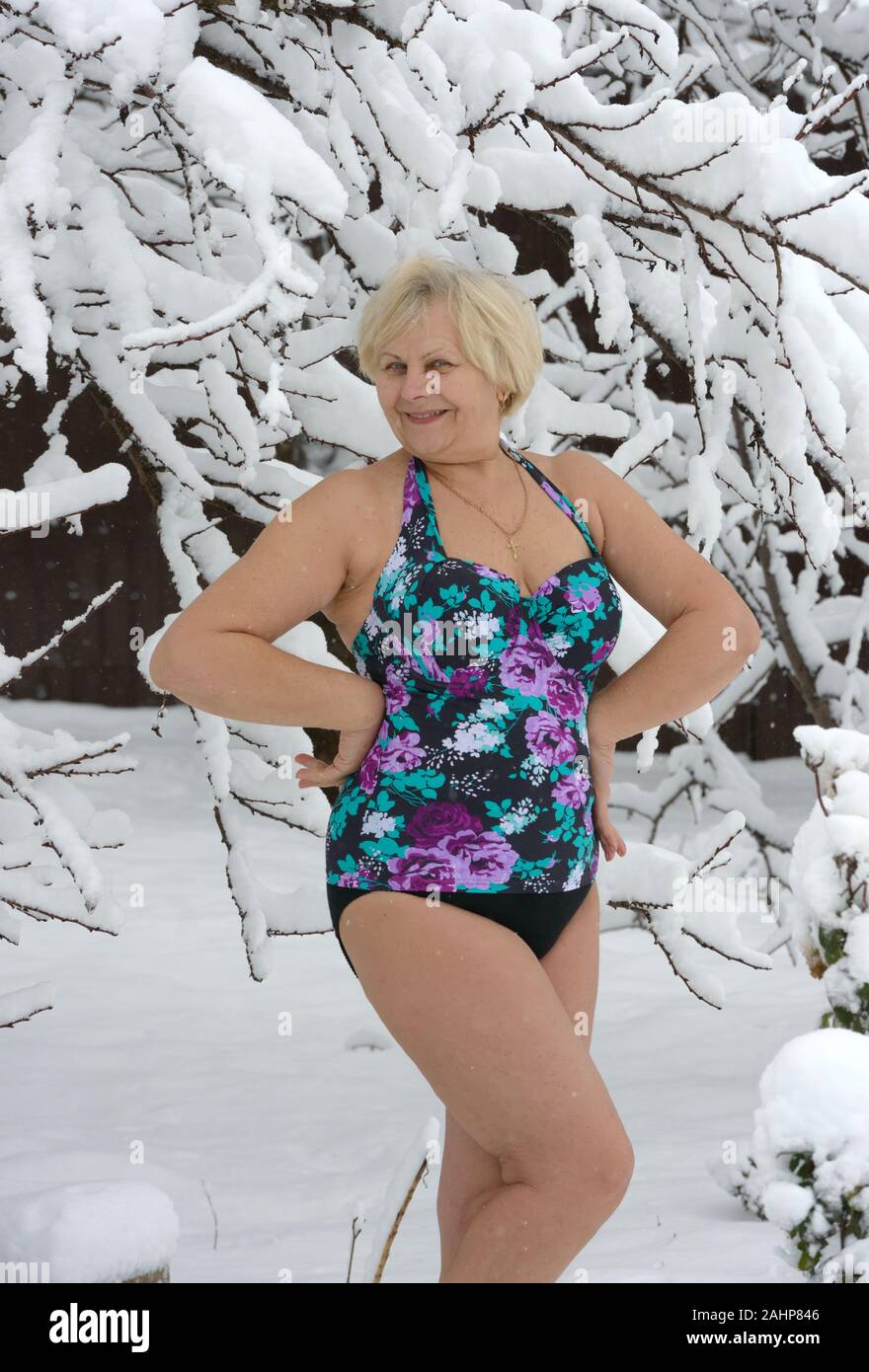 Close-up portrait of tanned smiling senior woman in beachwear that is posing near fresh snow-covered tree in the garden. Stock Photo