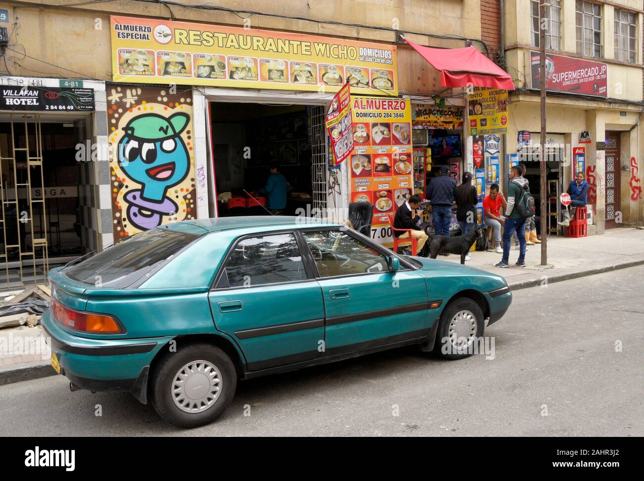 Small businesses, people socializing, and a car parked on Calle 18 in La Candelaria district of Bogota, Colombia Stock Photo