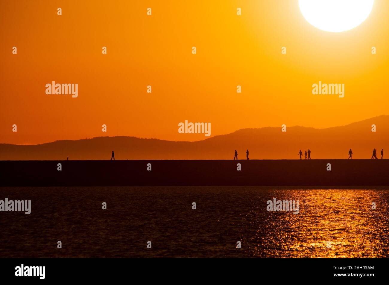 Silhouetted walkers on a jetty at the moment of a golden sunset, with sea in foreground and mountains in background Stock Photo
