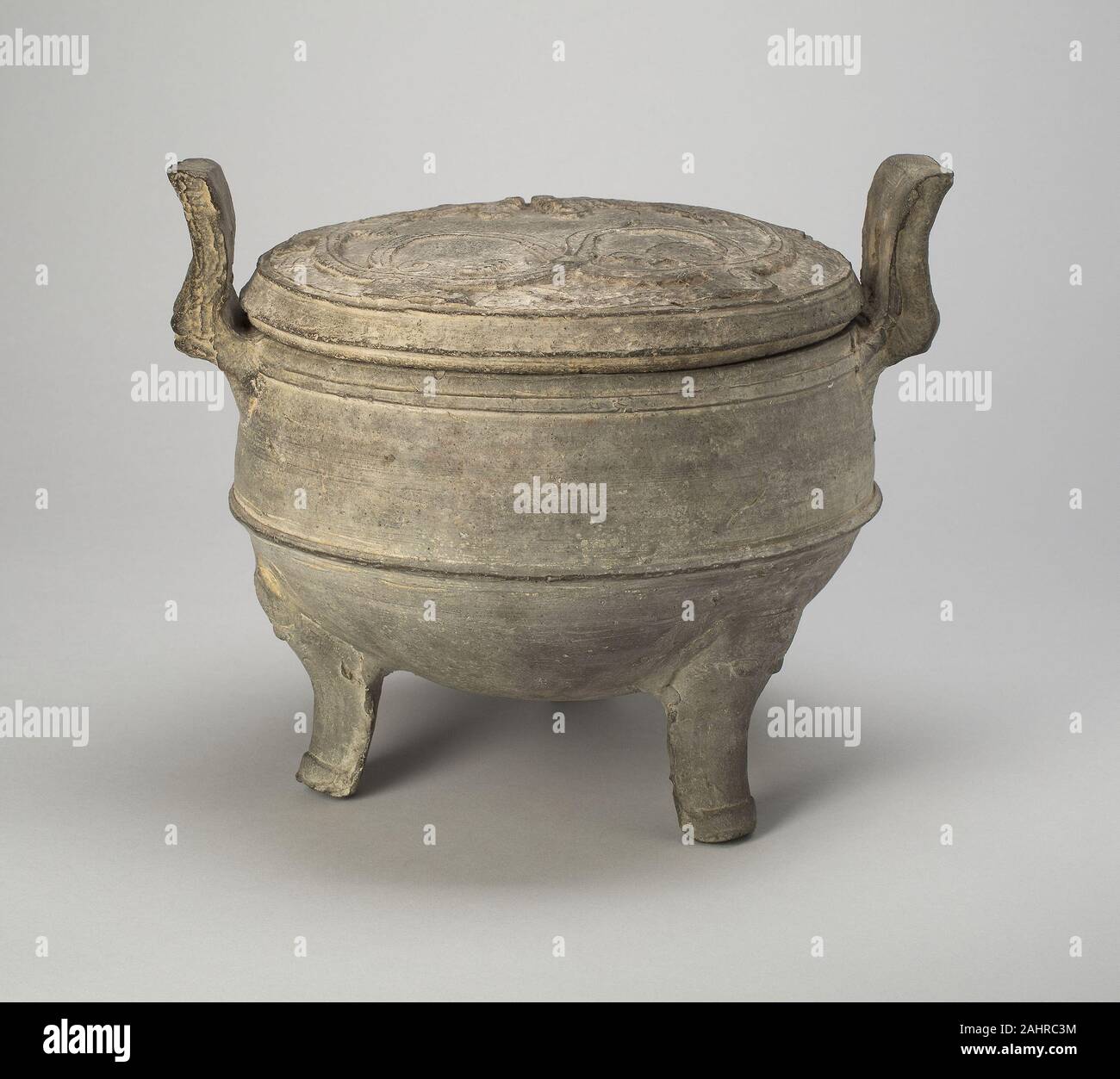 Tripod Caldron (Ding). 206 BC–9 AD. China. Gray earthenware with mold-impressed decoration Under the Han dynasty (206 B.C.-A.D. 220), dynastic stability and affluence broadened the clientele for burial goods from aristocratic families to officials and landowners. Clay replicas of costly vessels of bronze and lacquer met the wider demand for tomb furnishings by middle-income patrons. This caldron, modeled after a common bronze shape, is covered with a lid molded to depict a sinuous dragon—a design executed most naturally in lacquer painting. Stock Photo