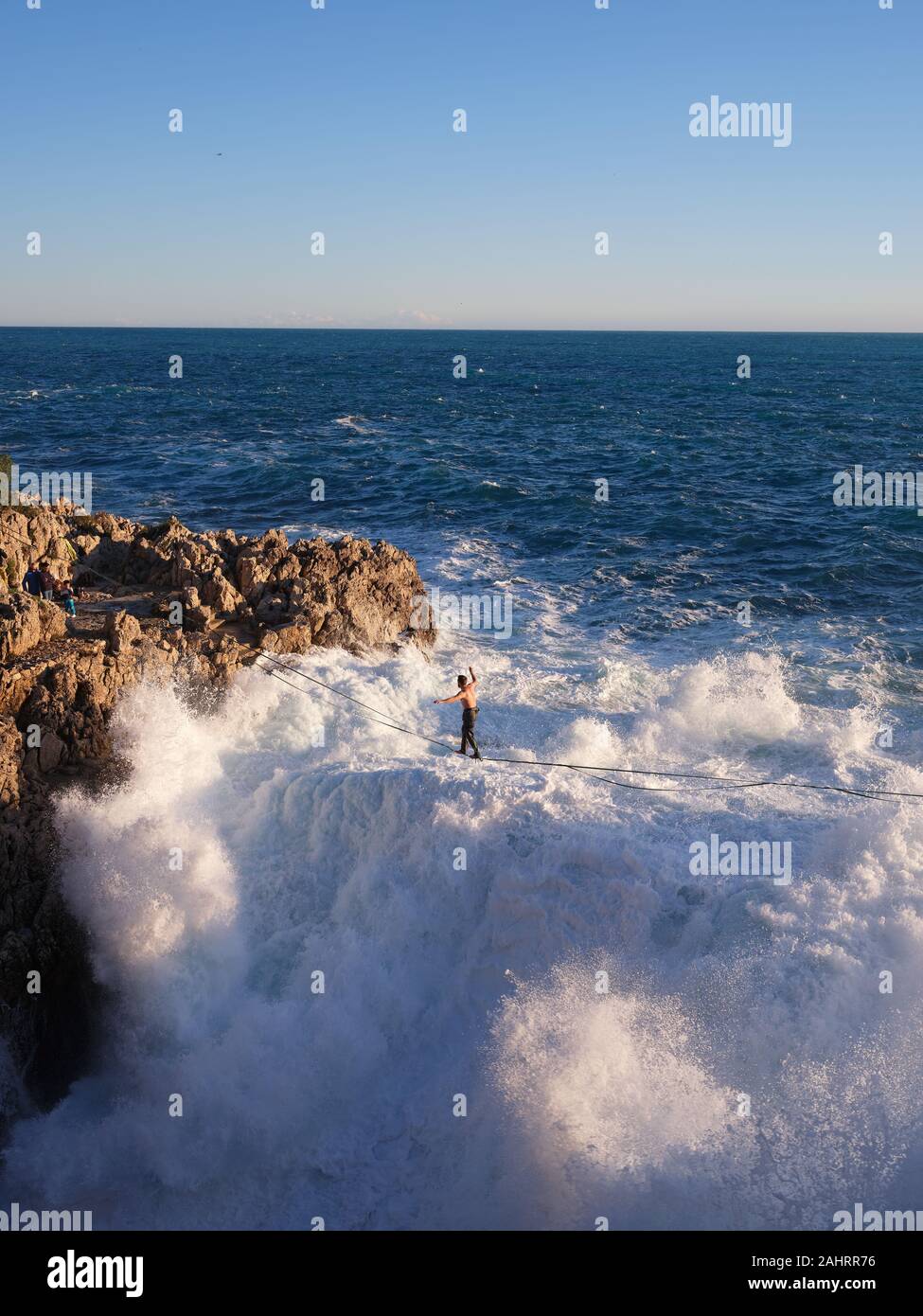 Young man slacklining above big waves crashing on a rocky coastline. Cap de Nice, French Riviera, Alpes-Maritimes, France. Stock Photo