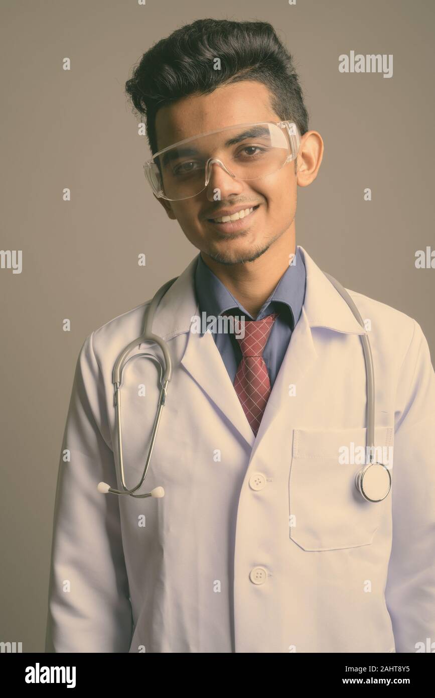 Studio shot of young Indian man doctor wearing protective glasses against gray background Stock Photo