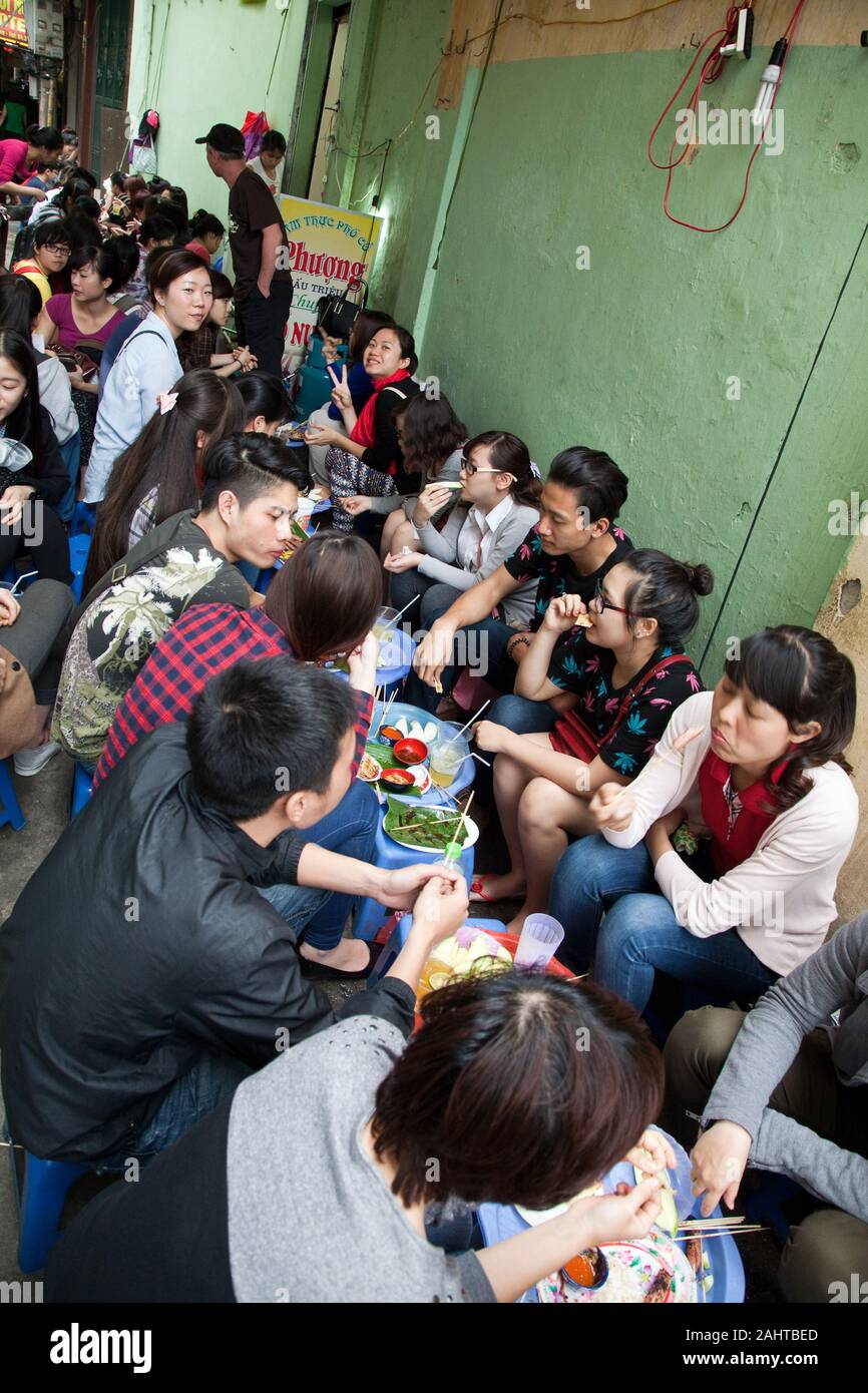 Group of young Asian people sitting on low plastic chairs eating their lunch or dinner together on the street Stock Photo