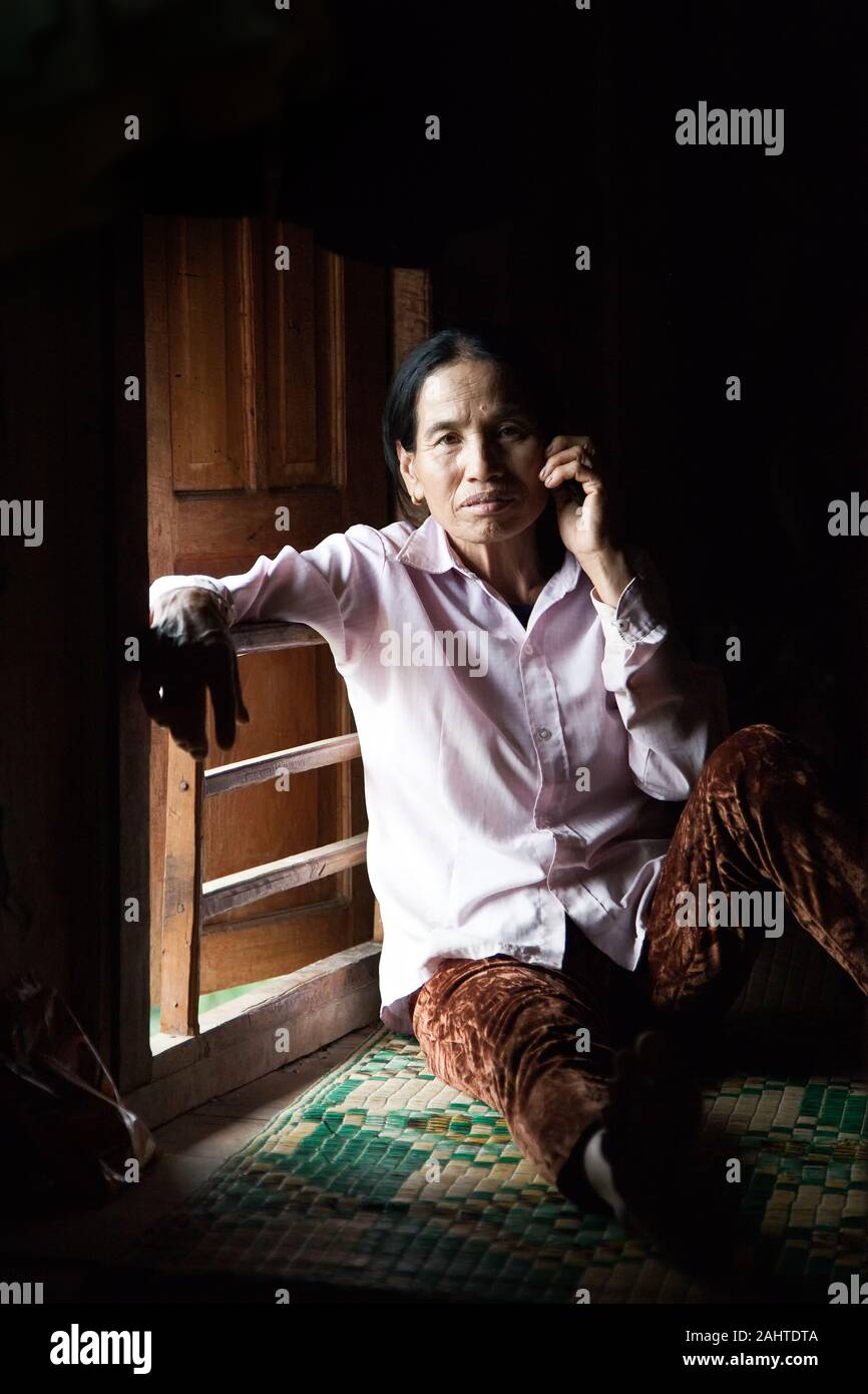Asian woman sits on the floor of her stilt house at the window opening where the sunlight enters Stock Photo