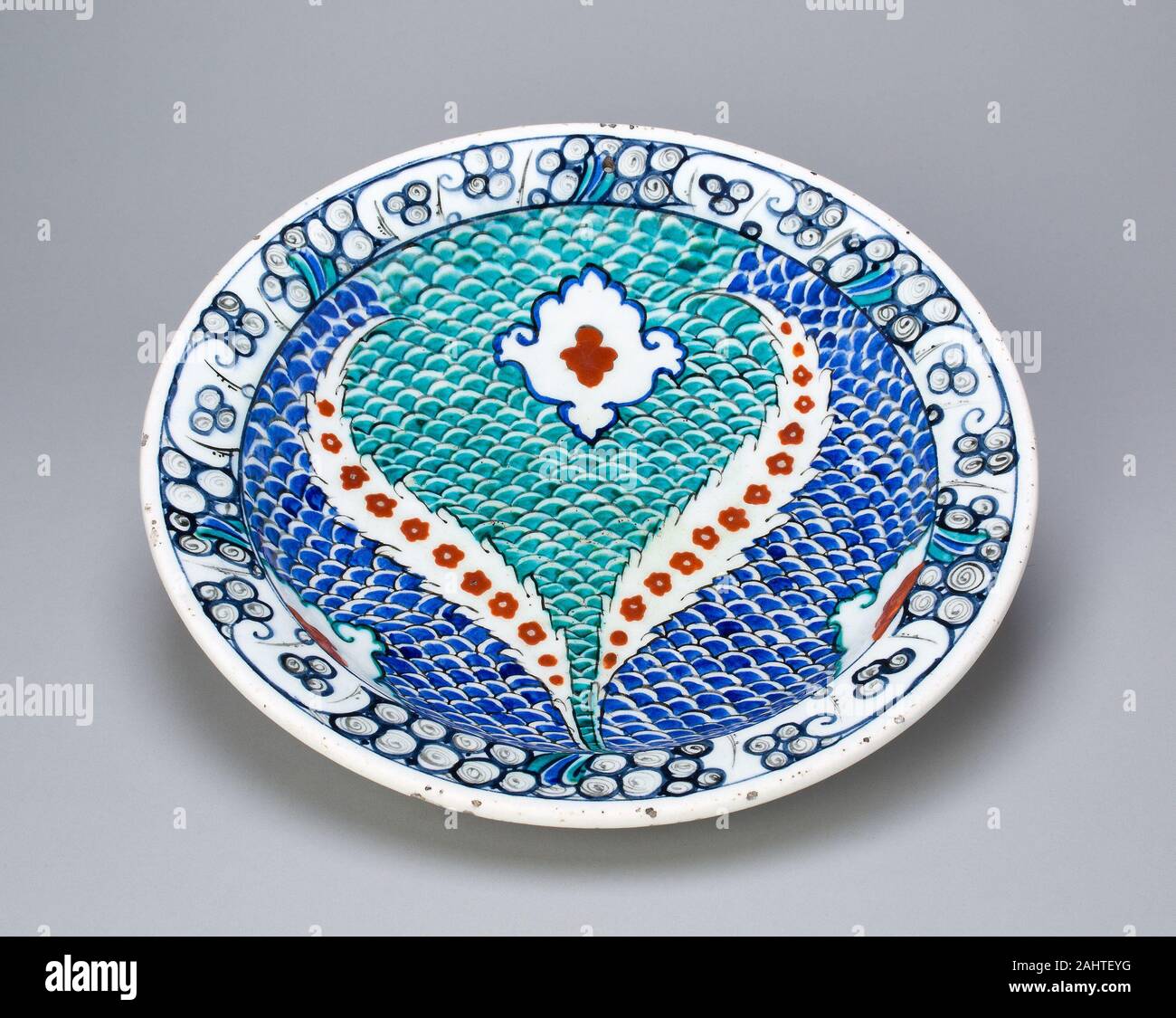 Islamic. Dish (Tabaq) with Scale Pattern and Serrated Leaves. 1550–1599. Turkey. Fritware with underglaze painting in blue, turquoise, red, and black The central design of this dish features two opposing leaves surrounding a central three-lobed (trefoil) shape, all set against a fish-scale motif in turquoise and blue. The design of the leaves is in the so-called saz (reed) style, referring to the reed pen used by Persian and Turkish artists to create black-ink drawings. Early drawings in this style depict dragons and phoenixes in combat surrounded by blossoms and feather-like leaves. The rim o Stock Photo