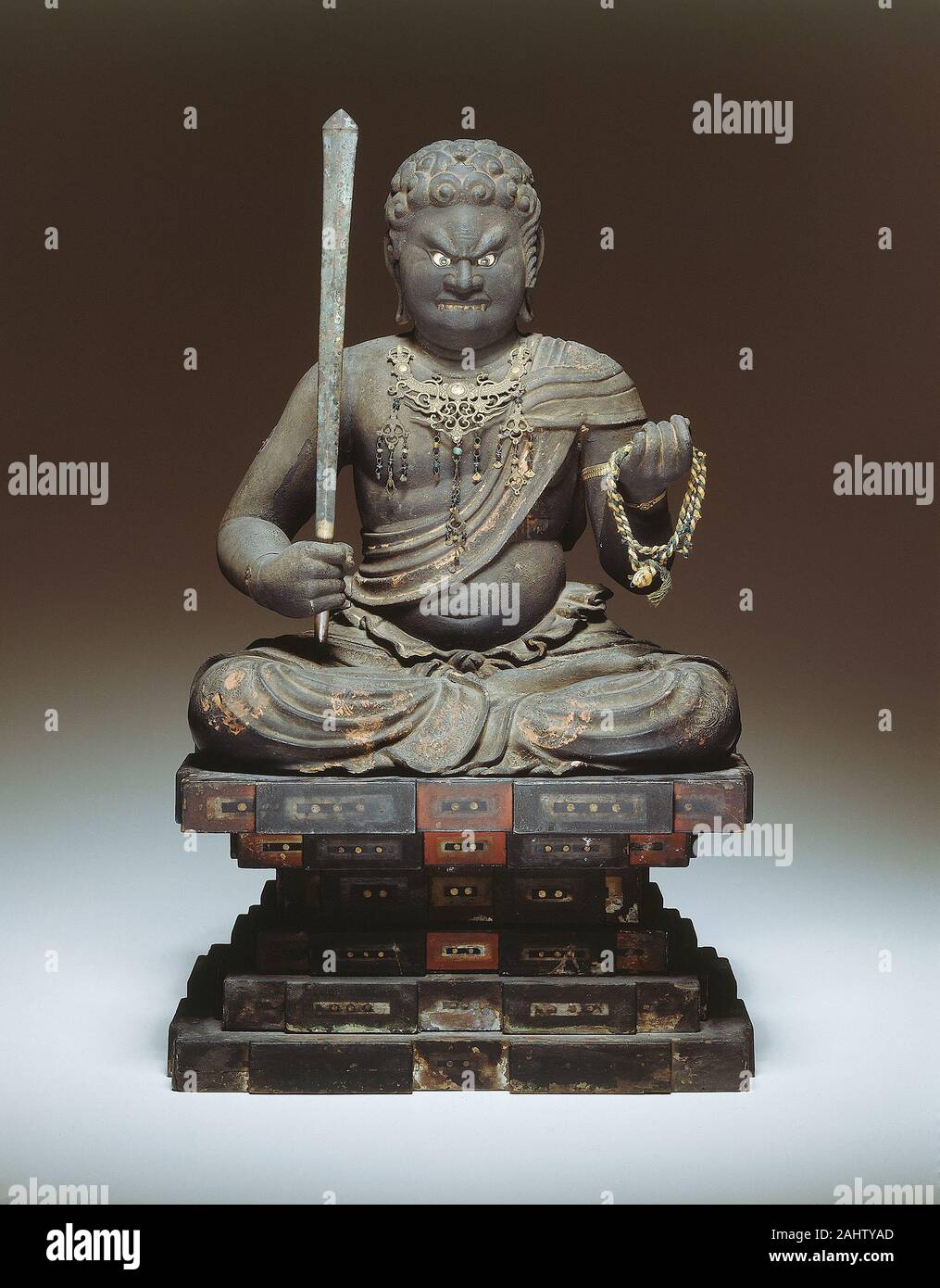 Fudo Myo-o. 1199–1399. Japan. Wood with polychromy and gilt-bronze accessories The name Fudo Myo-o means “the immovable or unshakable one.” He is one of five myo-o, or lords of light, whose threatening appearance guards the Law of Buddhism. He is equipped to guide the spiritual traveler past temptation on the path to enlightenment. Fudo’s bulging eyes, piercing stare, and protruding fangs express the intensity of his wrath against evil. Seated on a stylized rock formation that symbolizes his steadfastness, he once held his attributes, a rope and sword (these have been removed for conservation) Stock Photo