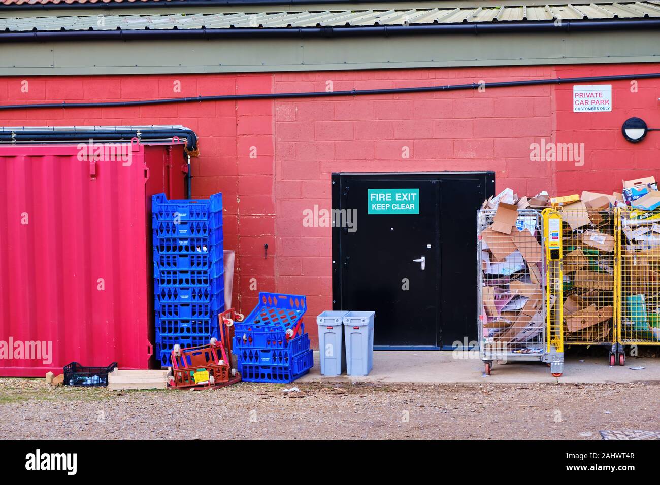 Black fire exit door in red painted building wall with sign keep clear surrounded by waste card and stacked boxes bins trash cans Stock Photo