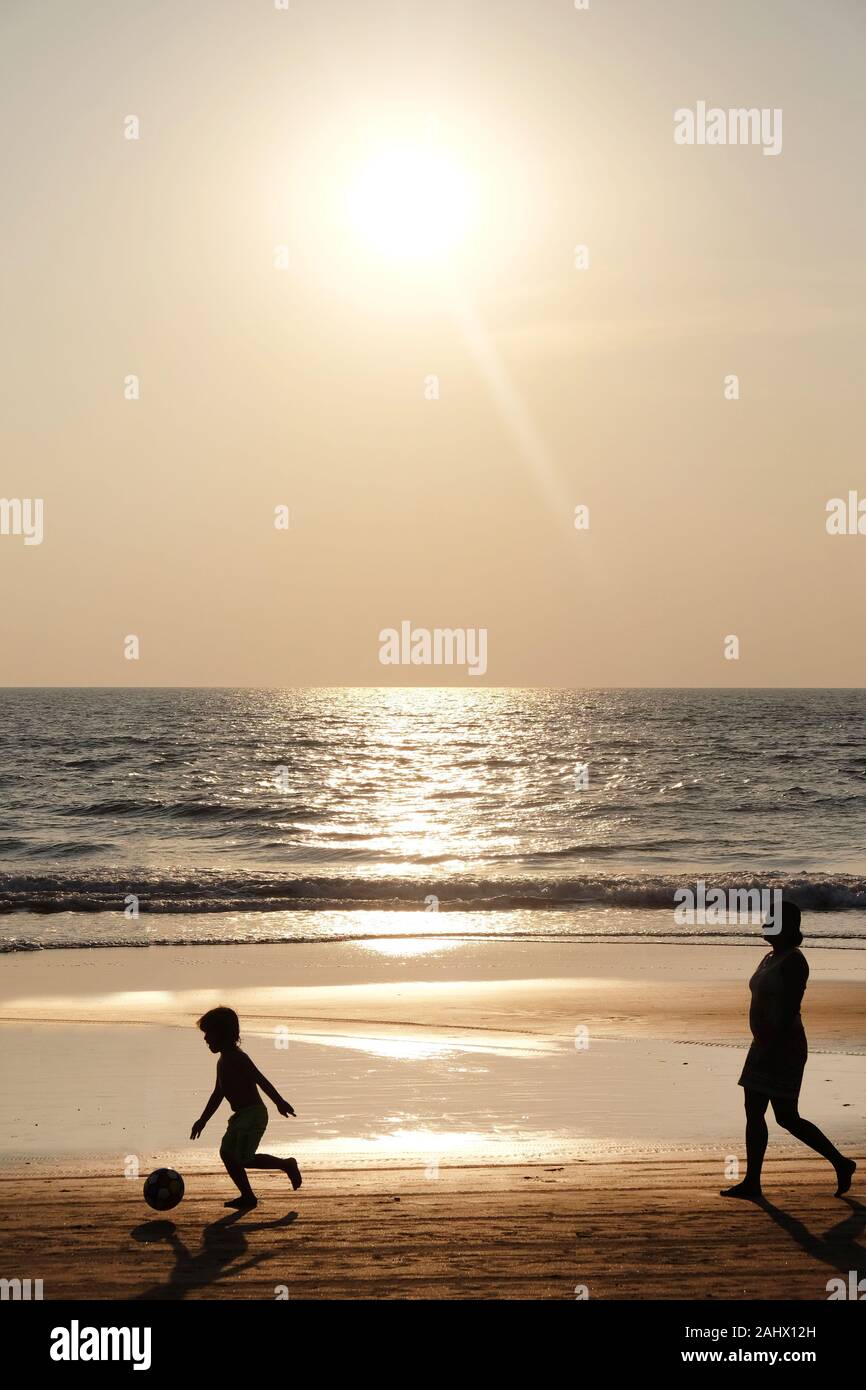 Silhouette of an unrecognizable small child holding a football, followed by his mom, they are both walking along an empty golden sandy beach at sunset Stock Photo