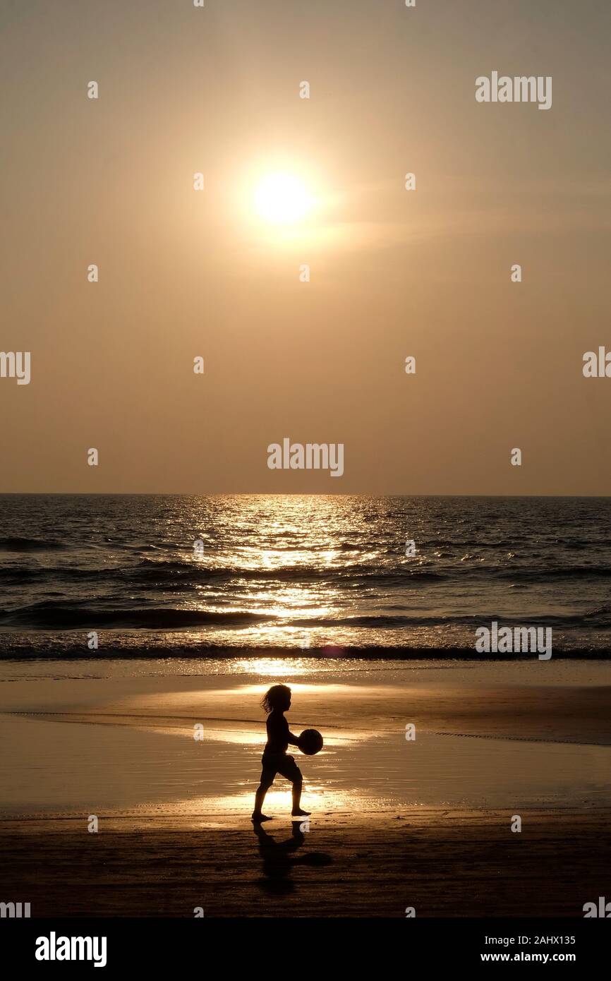 Silhouette of an unrecognizable small child holding a football walking along an empty golden sandy beach at sunset, a calm sea and clear golden sun li Stock Photo