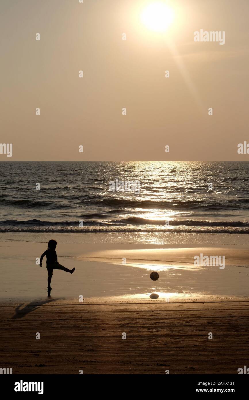 Silhouette of an unrecognizable small child kicking a football along an empty golden sandy beach at sunset, a calm sea and clear golden sun lit sky Stock Photo