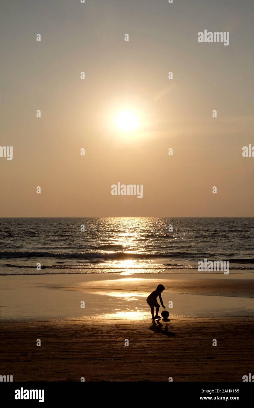 Silhouette of an unrecognizable small child picking up a football walking along an empty golden sandy beach at sunset, a calm sea and clear golden sun Stock Photo