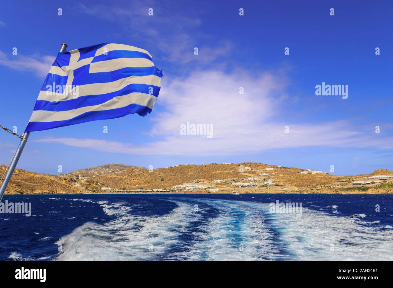 Discovering the Greek islands. Horizon with ship wake and the Greek flag: island of Mykonos in the far. Stock Photo