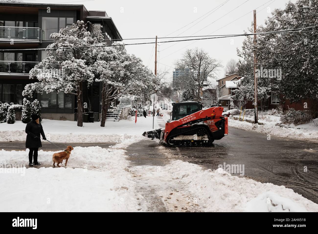 A woman walking a dog stop to watch a small, tracked Bobcat snowplow