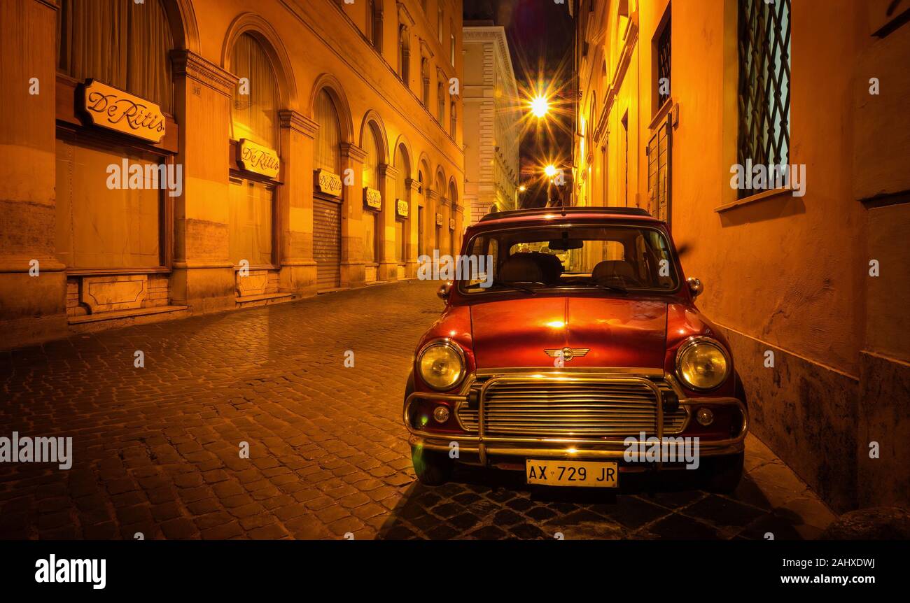 Rome, Italy - June 18, 2014. A red British Mini car parked on the side of a traditional cobblestone street late at night, under amber street lights. Stock Photo