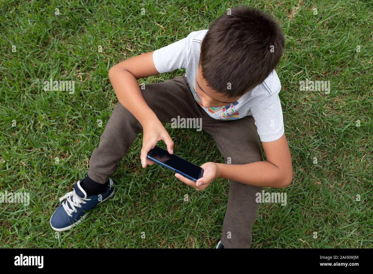 Close up of a little boy holding mobile phone in both hands while leaning arms on the knees. Concept of dependence and education on the use of Stock Photo