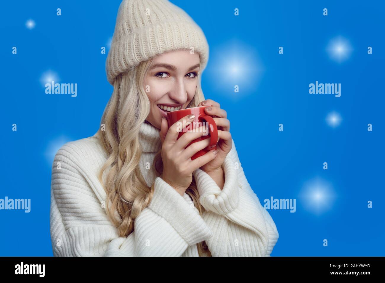 Young woman enjoying a mug of hot coffee in winter cupping it in her hands as she drinks with a blissful smile against a bluish background Stock Photo