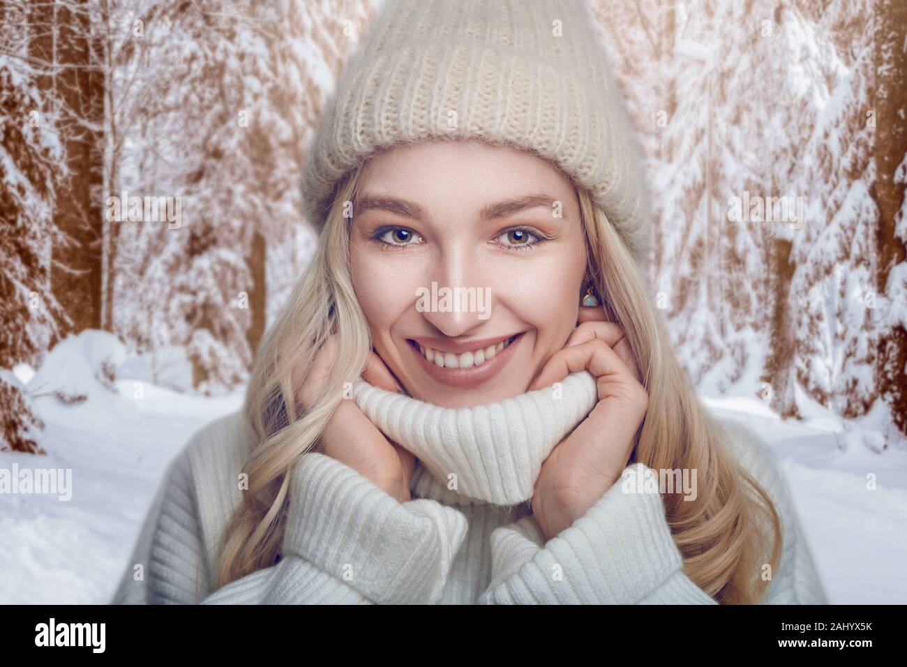 Attractive blond woman in a warm woollen polo neck sweater and knitted cap standing outdoors snuggling into the collar with a wide happy friendly smil Stock Photo