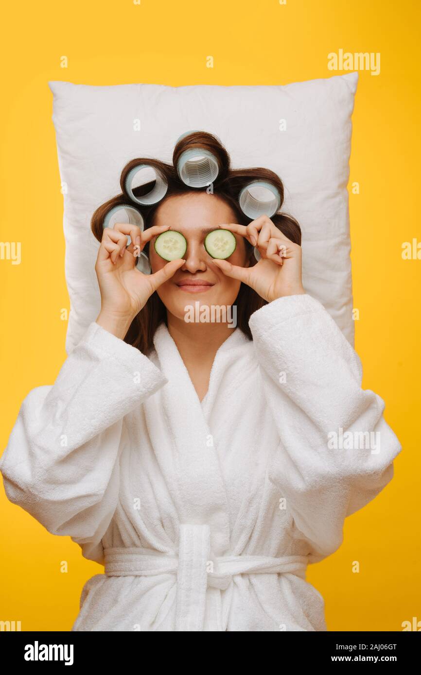 Woman lies on pillow with hair in rollers holding cucumber slices over her eyes Stock Photo