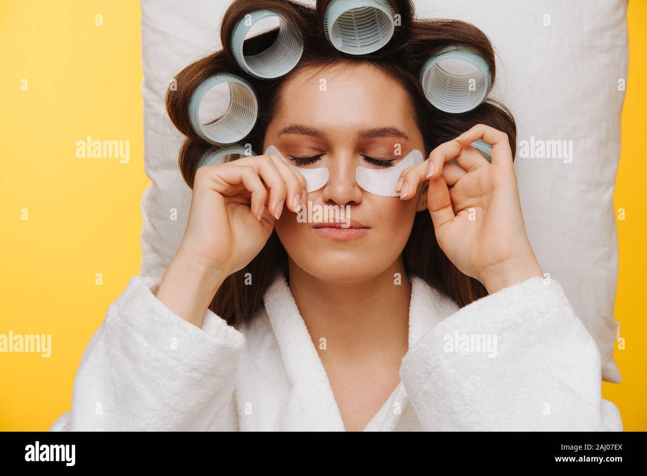 Woman lies on pillow with hair in rollers pressing patches under eyes