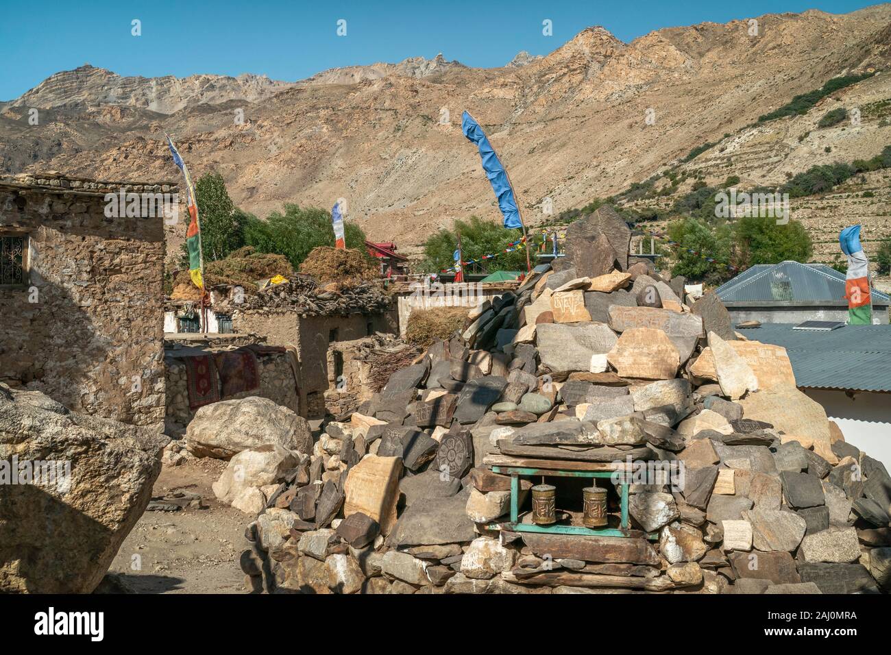 Street flanked by religious symbols, houses, prayer flags, and stone boulder, with Himalayas as backdrop on sunny day in Nako, Himachal Pradesh, India. Stock Photo