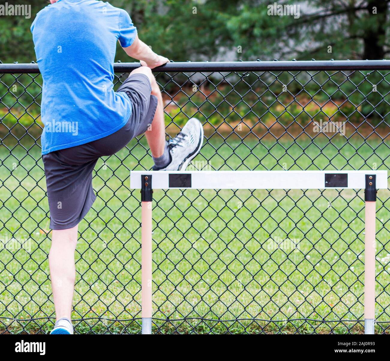 Rear view of a high school track and field athlete is performing a hurdle drill using a fence to balance at practice. Stock Photo