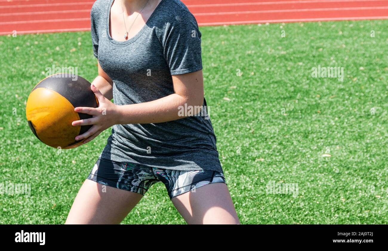 A high school girl is performing a core strength workout with a medicine ball  standing on a green turf field during practice. Stock Photo