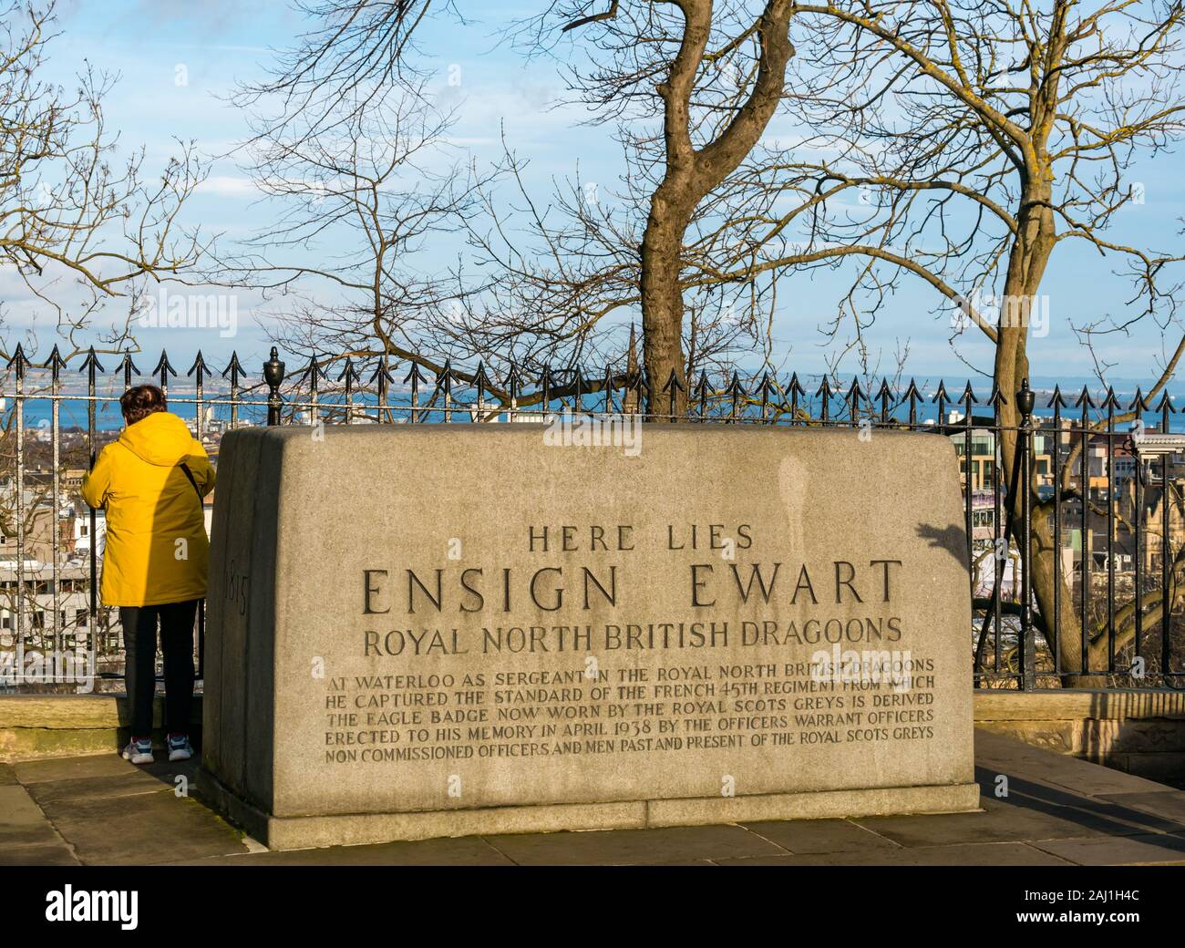 Female tourist wearing bright yellow coat admiring view by Ensign Ewart memorial, Edinburgh Castle esplanade, Edinburgh, Scotland, UK Stock Photo