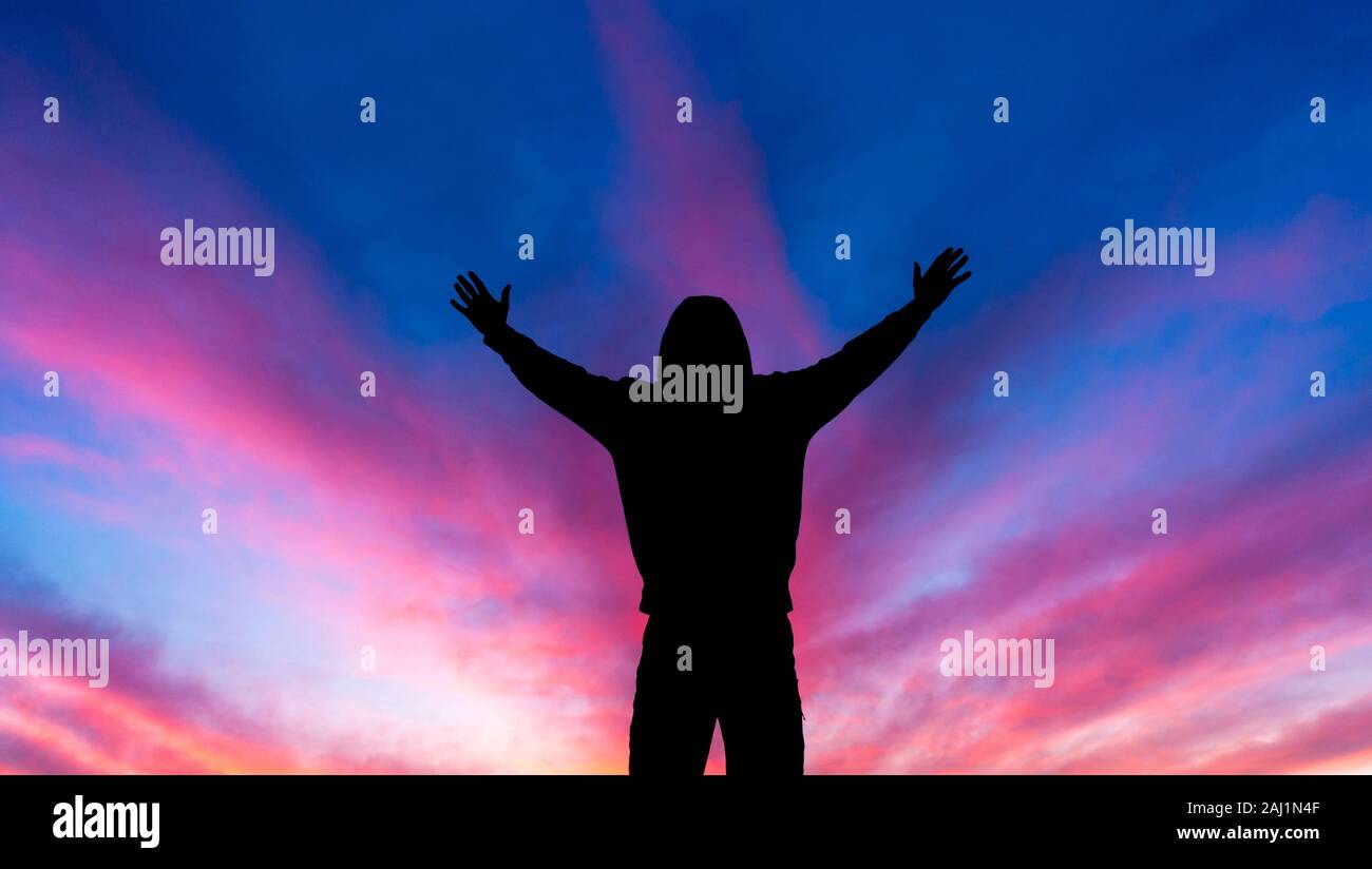 Rear view of man with arms outstretched against a colourful sky at sunset Stock Photo