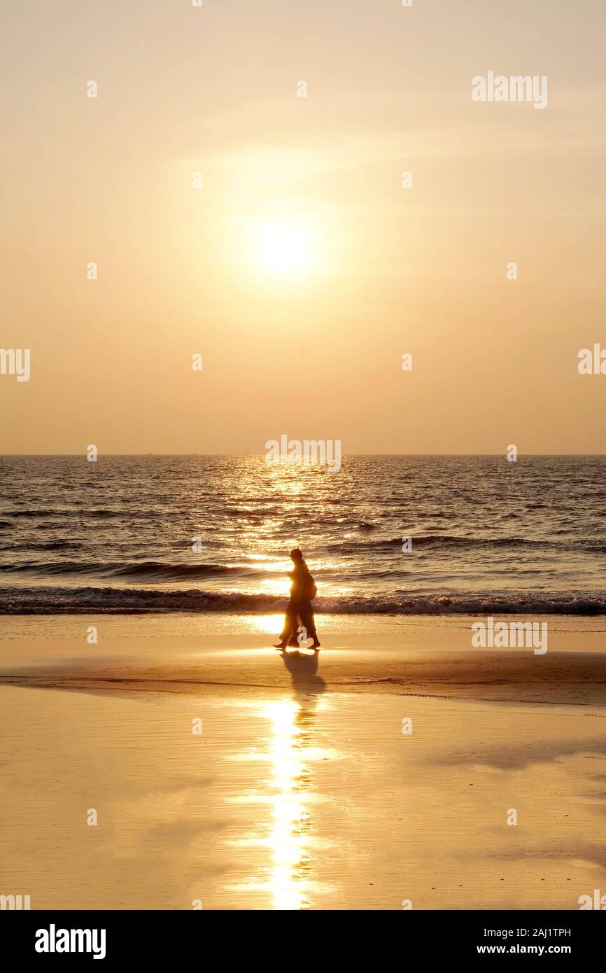 Silhouette of an unrecognizable woman wearing a traditional indian saris walking along an empty golden sandy beach at sunset, a calm sea and clear gol Stock Photo
