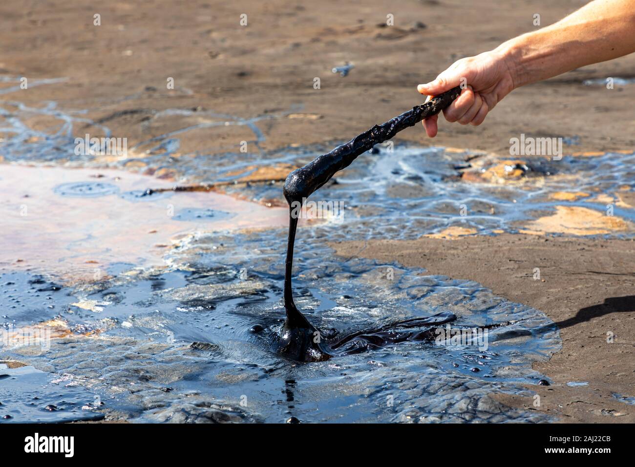 A man's hand stirring liquid asphalt with a wooden stick at Pitch Lake
