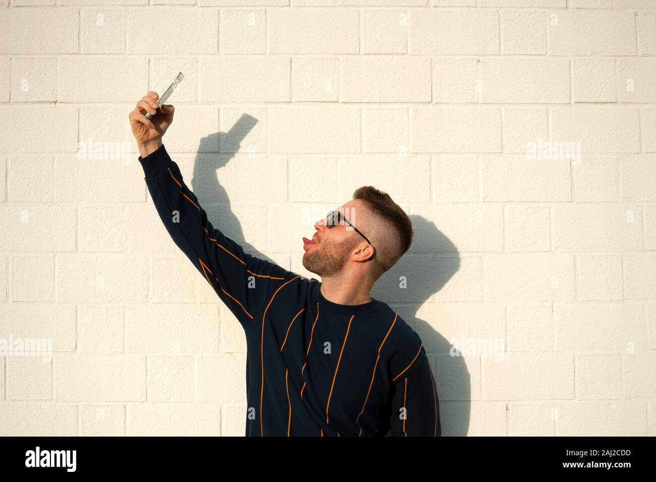 Positive smiling guy in casualwear showing tongue with mobile phone taking selfie. Young bearded man standing at outdoor white wall. Male selfie, leis Stock Photo