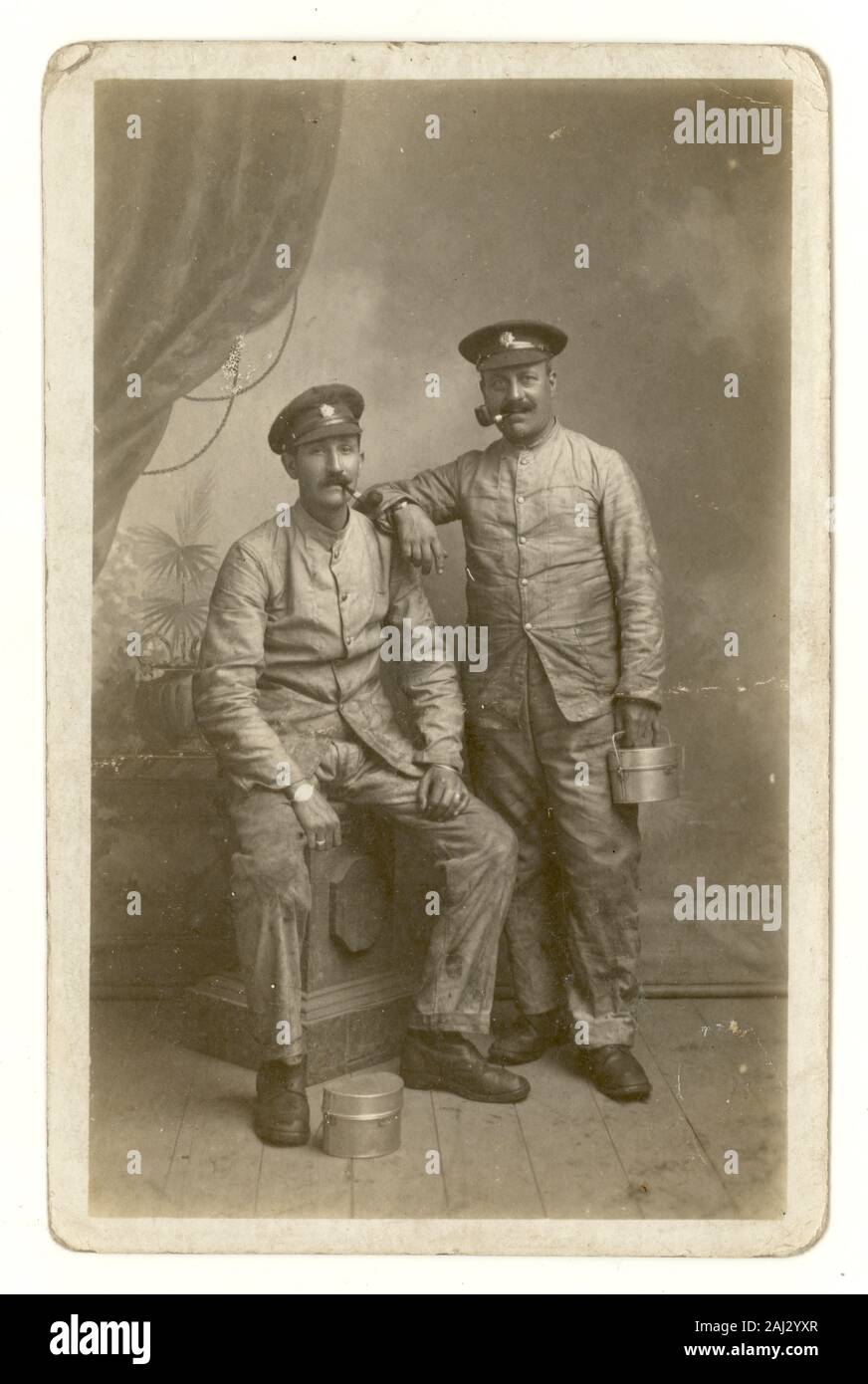 Original WW1 era portrait postcard of two cheerful men in the British Army Service Corps (ASC) at the Front in France. The friends are wearing fatigues and caps wearing the ASC badge, and carry a mess tin which may indicate they are soldier regimental cooks working in a field kitchen. Both men are smoking a pipe. U.K. date on reverse is 27 November 1917 and was sent from the Front in France. Stock Photo